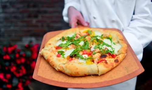 A chef holds a freshly baked pizza topped with vegetables on a wooden pizza peel, with flowers in the background.