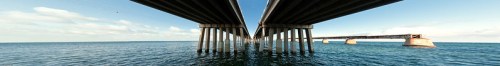 A panoramic view of two bridges spanning over calm water, supported by tall pillars under a clear blue sky.