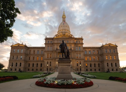 The Michigan State Capitol building at sunset, featuring a statue in the foreground and vibrant flower beds.