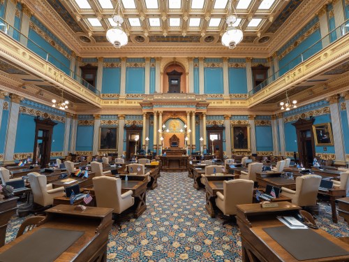 Interior of a grand legislative chamber with blue walls, ornate decor, and rows of desks, each with a small flag.