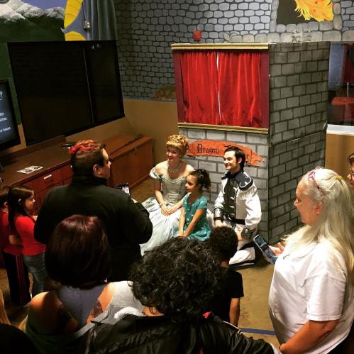 Children and adults gather around a puppet show featuring characters in a whimsical setting.