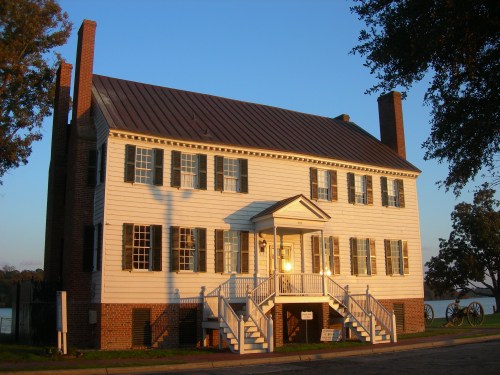 Historic two-story house with white siding, green shutters, and a metal roof, illuminated by sunset.