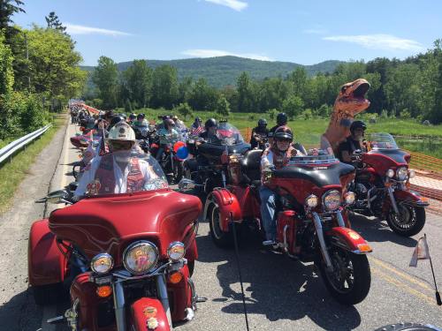 A parade of motorcycles, mostly red, with riders in helmets, including one in a dinosaur costume, against a scenic backdrop.