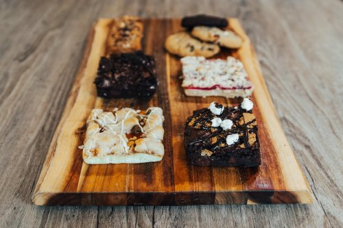 A wooden board displaying an assortment of baked goods, including brownies, cookies, and dessert bars.