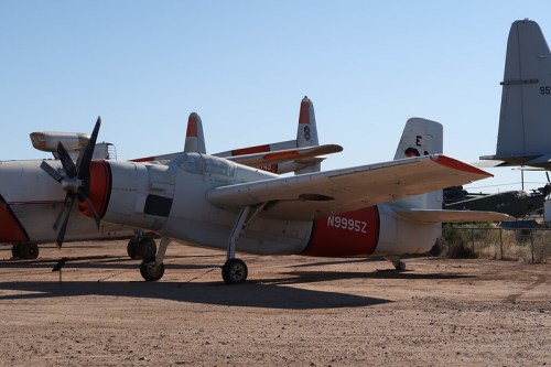 A vintage aircraft with a red and white color scheme parked on a dirt lot, surrounded by other planes in the background.