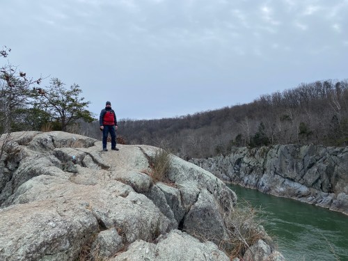 A person stands on a rocky outcrop overlooking a river, surrounded by trees and a cloudy sky.