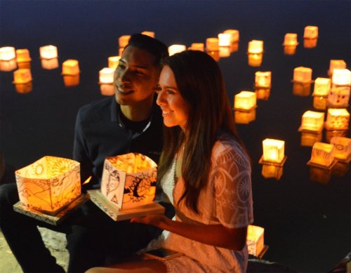 A young couple smiles while holding illuminated lanterns by a serene water surface, surrounded by glowing lanterns.