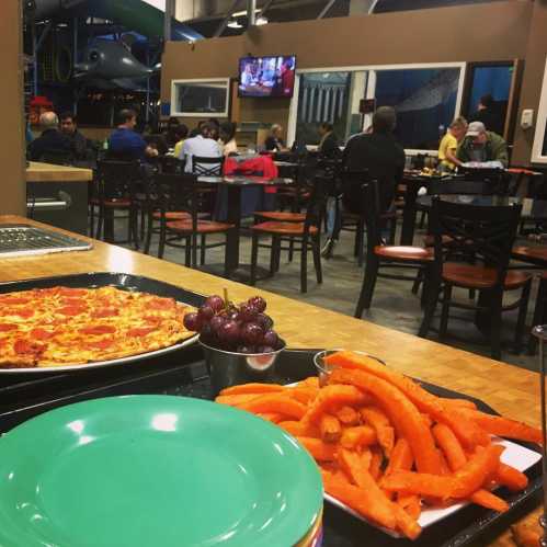 A plate with pizza, grapes, and sweet potato fries in a busy dining area with people seated in the background.