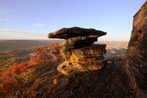 A large, flat rock balanced on top of smaller rocks, surrounded by autumn foliage and a scenic valley view.