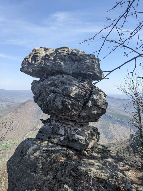 A large, balanced rock formation atop a cliff, with a scenic view of rolling hills and a clear blue sky in the background.
