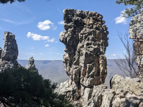 A rocky outcrop stands tall against a blue sky, surrounded by mountains and sparse trees.