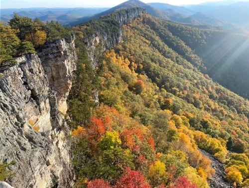 A scenic view of a mountain range with vibrant autumn foliage and rocky cliffs under a clear blue sky.