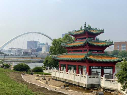 A traditional Chinese pavilion in a park, with a modern city skyline and arch bridge in the background.