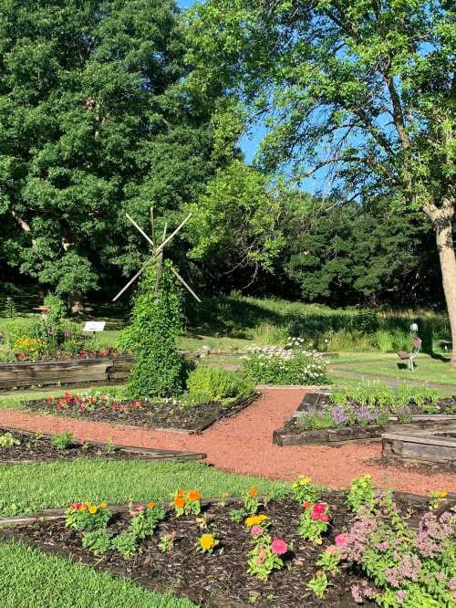 A vibrant garden with flower beds, a wooden windmill, and lush greenery under a clear blue sky.