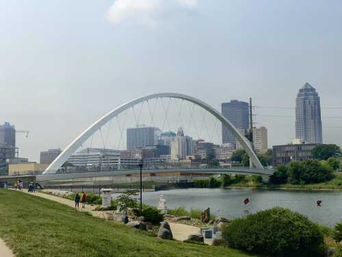 A modern arch bridge spans a river with a city skyline in the background, featuring tall buildings and greenery along the shore.