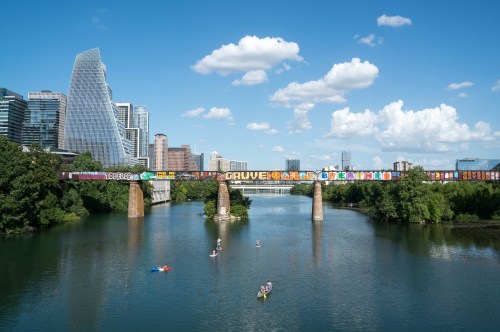 A vibrant cityscape with a bridge covered in graffiti, over a calm river with people kayaking under a blue sky.