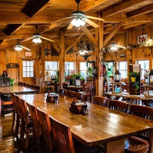 A cozy wooden dining area with large tables, ceiling fans, and rustic decor, surrounded by plants and warm lighting.