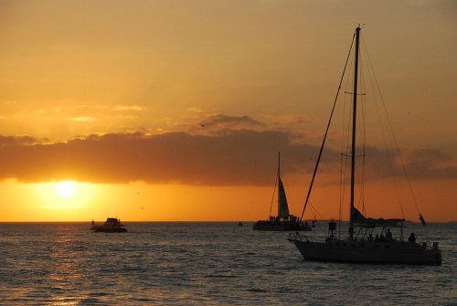 Sailboats on the water at sunset, with a vibrant orange sky and clouds reflecting the evening light.