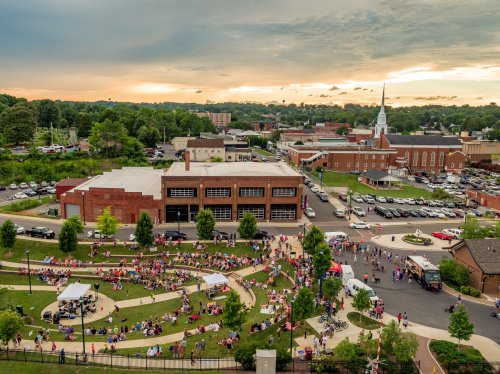 Aerial view of a crowd gathered in a park during an event, with buildings and a church in the background under a cloudy sky.
