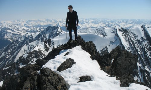 A person stands on a snowy mountain peak, overlooking a vast range of snow-capped mountains under a clear blue sky.