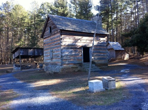 A rustic wooden cabin surrounded by trees, with a stone foundation and a nearby historical marker.
