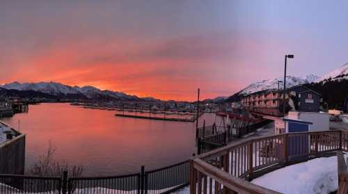 A panoramic view of a serene harbor at sunset, with mountains in the background and colorful skies reflecting on the water.