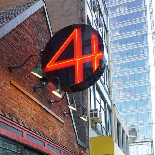 A round neon sign with the number "4" illuminated in red, mounted on a brick wall next to modern glass buildings.