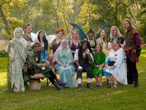 A diverse group of people dressed in fantasy costumes, including fairies and mythical characters, posing outdoors by a river.