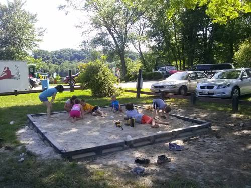 Children playing in a sandpit, digging and building, surrounded by trees and parked cars in a sunny outdoor setting.