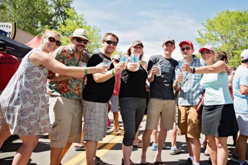 A group of eight people smiling and raising drinks together at an outdoor event on a sunny day.