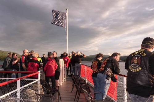 A group of people on a boat, enjoying the view, with a flag flying and mountains in the background.
