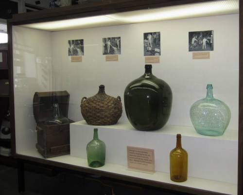 Display case featuring various glass bottles and a woven basket, with historical photos above.
