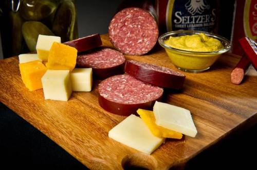 A wooden board with assorted cheeses, sliced salami, mustard, and pickles.