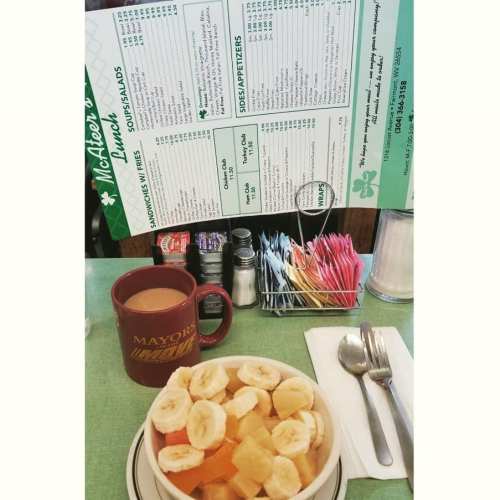 A bowl of fresh fruit with bananas and melons, a coffee mug, and a menu on a diner table.