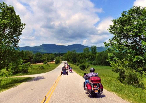 A line of motorcycles rides along a scenic road with green hills and trees under a partly cloudy sky.
