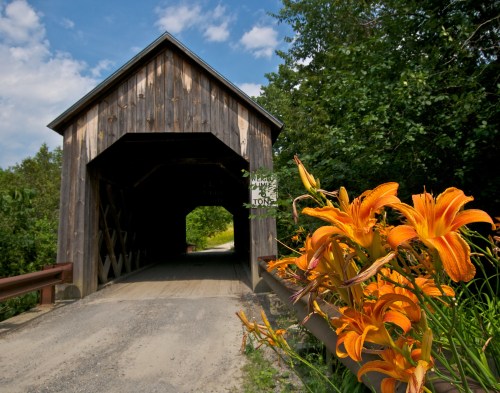 A wooden covered bridge with a gravel path, framed by vibrant orange lilies and lush green trees under a blue sky.