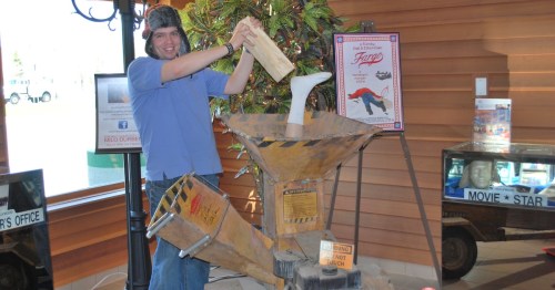 A person in a hat is playfully using a wooden plank on a large, industrial-looking machine in a casual setting.