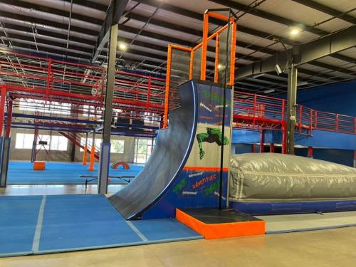 Indoor trampoline park featuring a curved ramp, foam pit, and various climbing structures. Brightly colored and spacious.