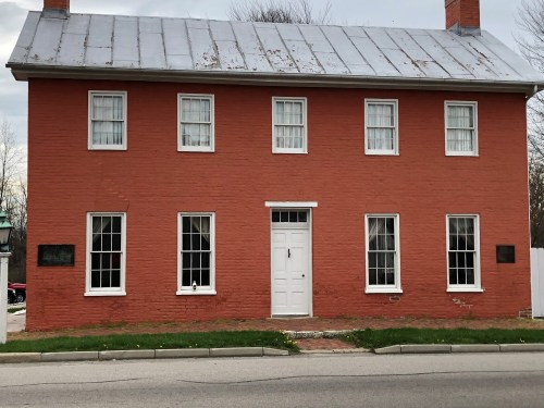 A two-story red brick house with a metal roof, featuring white windows and a central door, set on a grassy street.