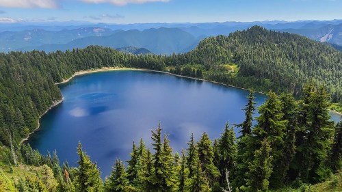 A serene mountain lake surrounded by lush green forests and distant blue mountains under a clear sky.