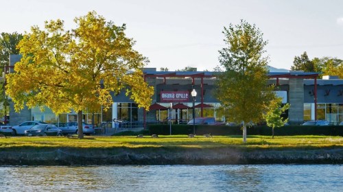A restaurant with a red sign, surrounded by trees, by a river on a sunny day.