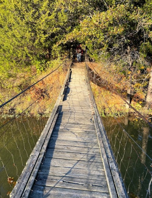A wooden suspension bridge stretches over a calm body of water, surrounded by trees and autumn foliage.
