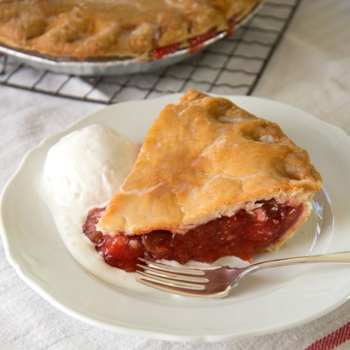 A slice of cherry pie on a white plate with a scoop of vanilla ice cream and a fork beside it.