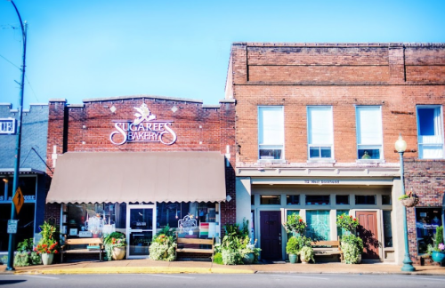 A charming bakery with a brown awning and flower-filled planters, set in a historic brick building on a sunny street.