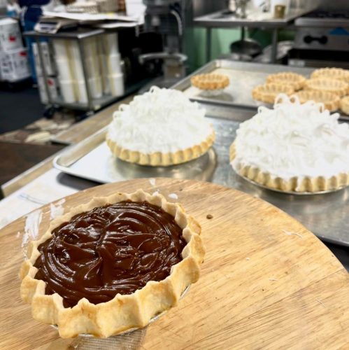 A wooden board with a chocolate pie and two meringue-topped pies in the background, set in a kitchen.