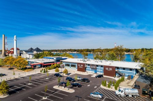 Aerial view of a waterfront building with a parking lot, surrounded by trees and a river under a clear blue sky.