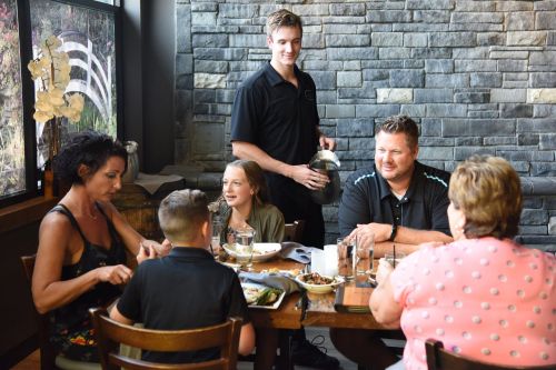 A family enjoys a meal at a restaurant, with a waiter serving drinks and dishes at their table.