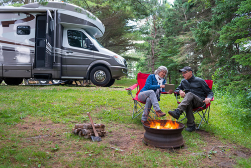A couple sits by a campfire in front of their RV, enjoying each other's company in a wooded area.