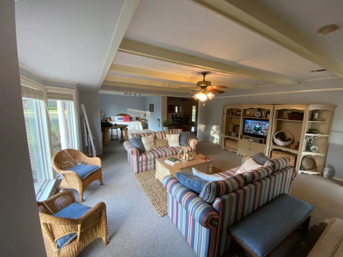 Cozy living room with striped sofas, wicker chairs, a coffee table, and a TV, featuring large windows and soft lighting.