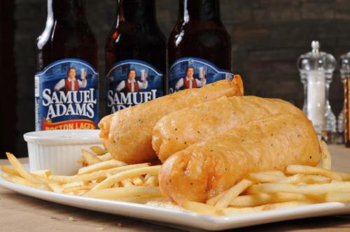 A plate of golden fried fish fillets served with French fries and three bottles of Samuel Adams beer in the background.